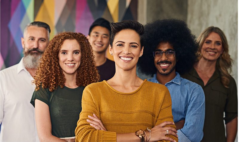 Six smiling people of diverse backgrounds stand together in front of a colorful, geometric-patterned wall; the woman in front wears a mustard sweater and has her arms crossed confidently.